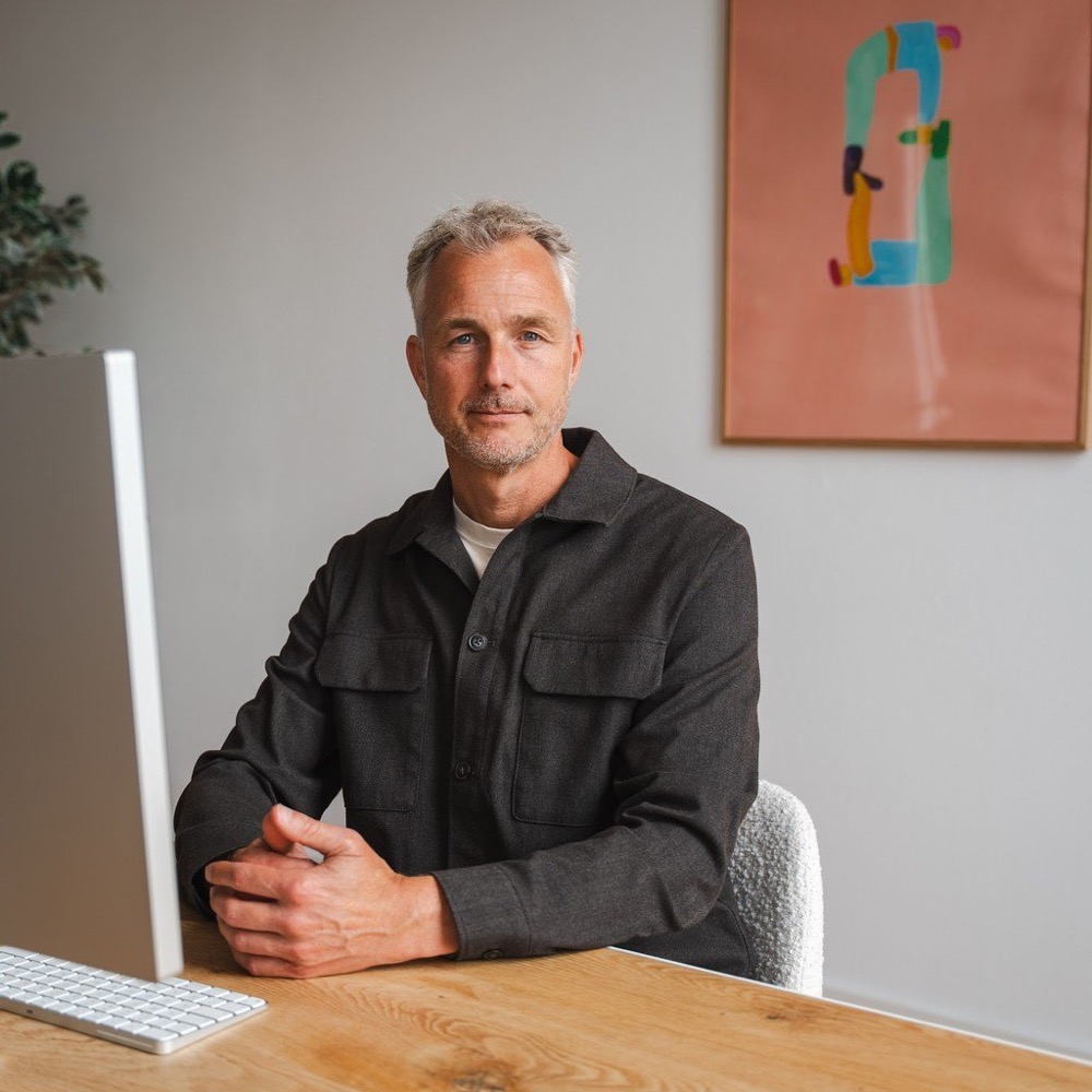Mischa Diender aan een eiken bureau met een computer, met een schilderij op de achtergrond.